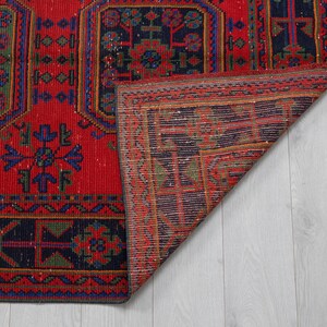 May include: A rectangular woven rug featuring a vibrant red background with dark blue and green geometric patterns. The rug displays repeating octagonal motifs and intricate border designs. One corner of the rug is folded back to reveal the underside.