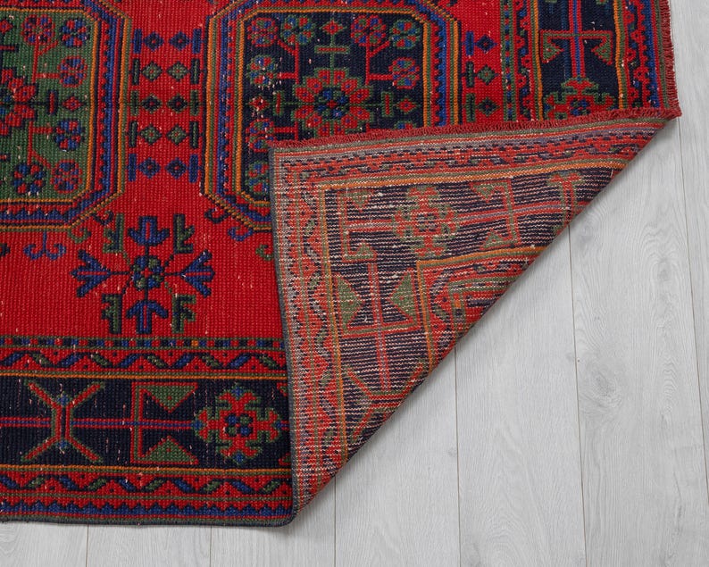 May include: A rectangular woven rug featuring a vibrant red background with dark blue and green geometric patterns. The rug displays repeating octagonal motifs and intricate border designs. One corner of the rug is folded back to reveal the underside.