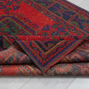 May include: A woven area rug featuring a vibrant red background with intricate geometric patterns in navy blue, forest green, and orange. The rug is folded over to reveal the detailed underside, showcasing a dense, textured pile and knotted construction.