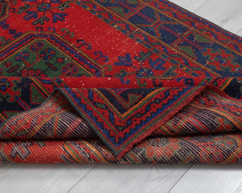 May include: A woven area rug featuring a vibrant red background with intricate geometric patterns in navy blue, forest green, and orange. The rug is folded over to reveal the detailed underside, showcasing a dense, textured pile and knotted construction.