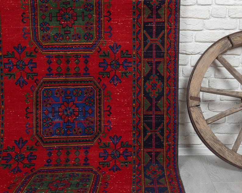 May include: A rectangular area rug featuring a vibrant red background with repeating geometric patterns in blue and green. The design includes central square motifs and a dark border with intricate linear details and floral-inspired accents.