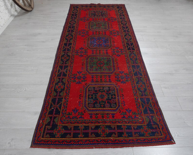 May include: A rectangular runner rug featuring a vibrant red field with five repeating geometric medallions in shades of blue and green. The border is dark blue with a repeating cross and diamond pattern, framed by thin red and blue lines.