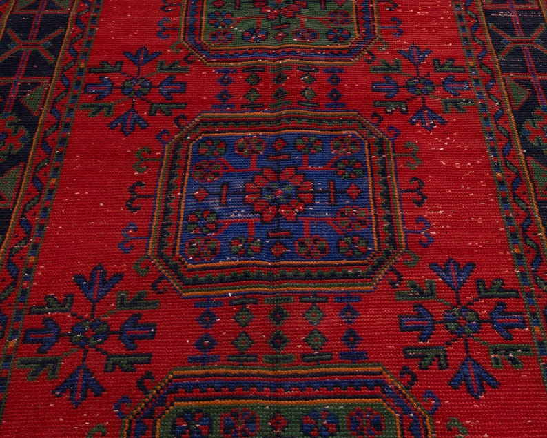 May include: A rectangular woven rug featuring a vibrant red background with repeating geometric patterns in blue and green. The design includes central octagonal motifs surrounded by stylized floral shapes and a dark blue border with geometric lines.