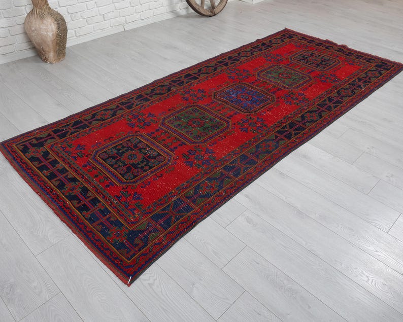 May include: A rectangular area rug featuring a vibrant red background with a series of repeating dark blue and green geometric medallions. The border consists of intricate dark blue patterns that frame the central design of the woven textile.