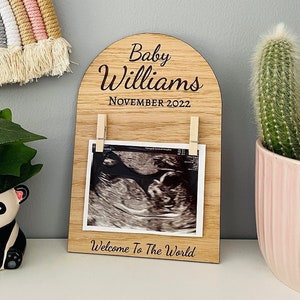 May include: Wooden photo display with an arched top, engraved with "Baby Williams November 2022". The display has two clothespins holding a black and white ultrasound image. The bottom of the display reads "Welcome to the World".