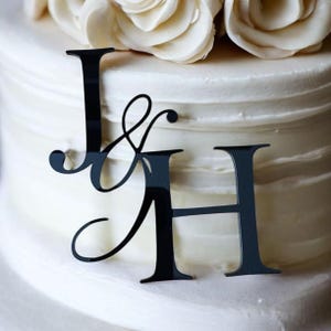 May include: Black acrylic cake topper with the letters "J & H" and an ampersand. The topper sits on a white frosted cake, with white flowers in the background. A decorative element for weddings or anniversaries.