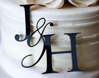 May include: Black acrylic cake topper featuring the letters "J & H" with an ampersand symbol. The topper is placed on a white frosted cake, with white flowers in the background. A decorative element for weddings or anniversaries.