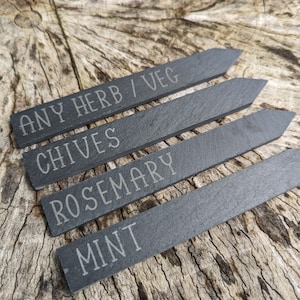 May include: Four black slate plant markers with white engraved text. The markers read "ANY HERB/VEG", "CHIVES", "ROSEMARY", and "MINT".