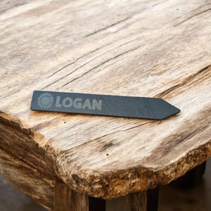 May include: A black slate garden marker with the word "LOGAN" engraved on it. The marker is shaped like an arrow and is sitting on a wooden surface.