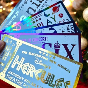 May include: A close-up of a blue ticket with gold foil lettering that reads "Disney's New Musical Hercules" and "Saturday 5th July 2020 Theatre Row".