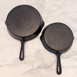 May include: Two black cast iron fry pans with handles. The larger pan is marked No 9 and the smaller pan is marked No 8. Both pans feature a shield logo with the letters GSW and the text FRY PAN and MADE IN CANADA embossed on the bottom surface.