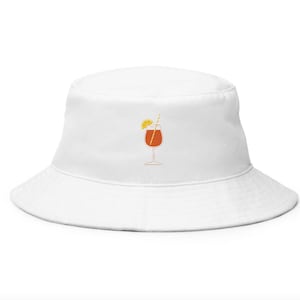 May include: White bucket hat with an embroidered orange cocktail design. The cocktail illustration includes a glass, a straw, and a lemon slice. The hat is a casual accessory, perfect for sunny days and outdoor events.