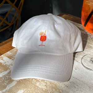 Pode incluir: Bon&eacute; de beisebol branco com um design de coquetel bordado em laranja e vermelho.
