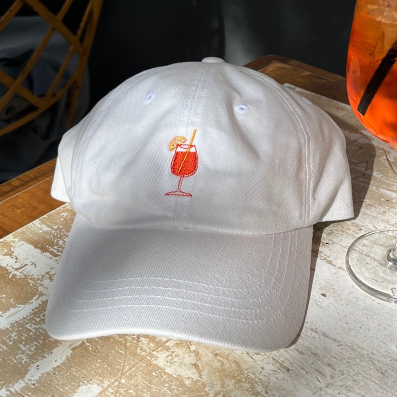 Pode incluir: Bon&eacute; de beisebol branco com um design de coquetel bordado em laranja e vermelho.