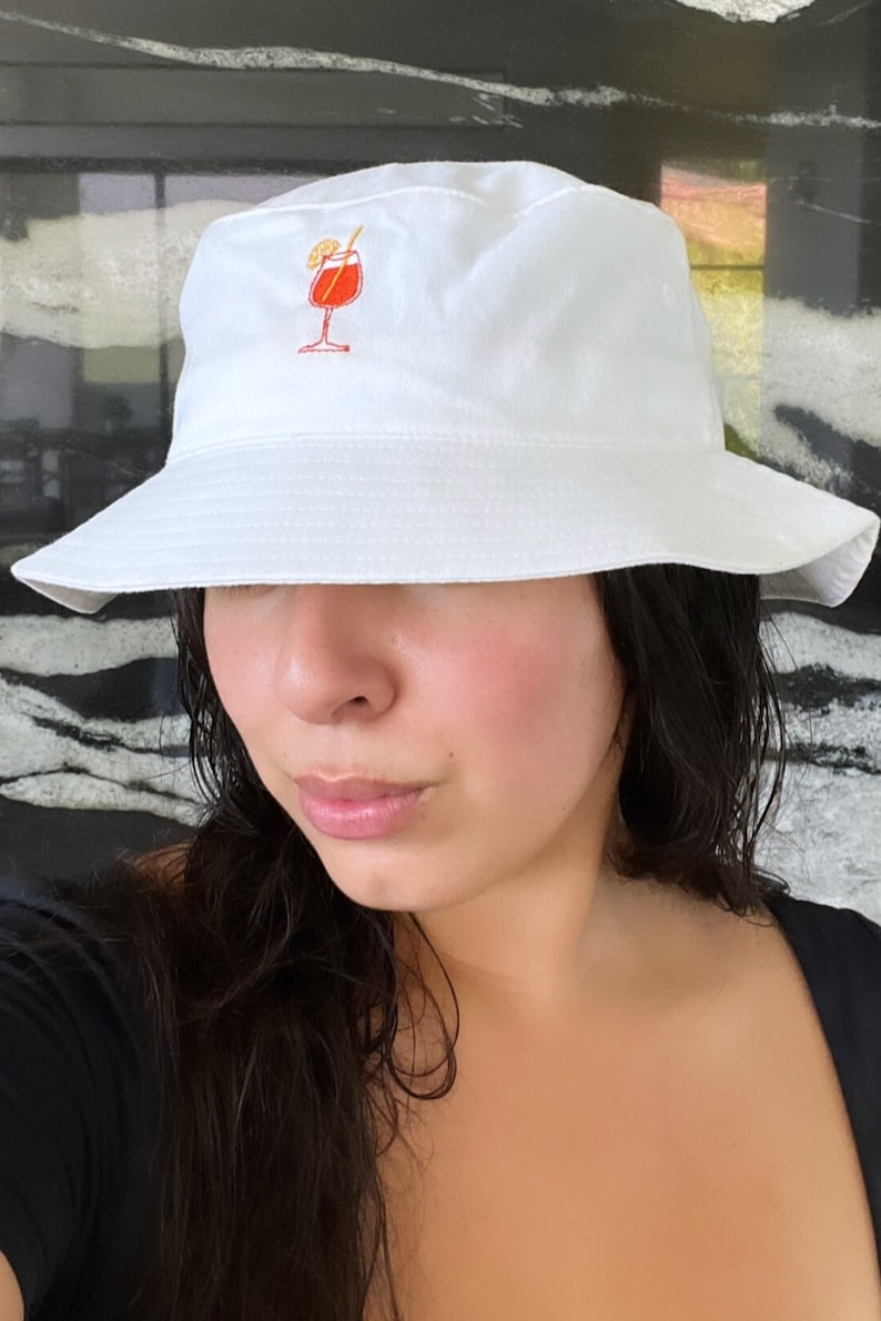 May include: White bucket hat with a small embroidered cocktail design in red and orange. The hat has a wide brim and is made of a soft fabric. A casual accessory for sunny days.