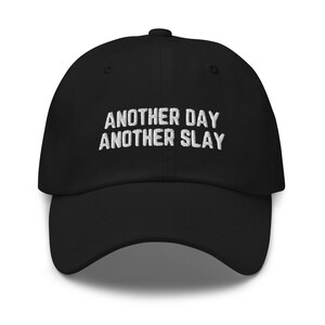 Op de afbeelding: Zwarte baseballpet met witte geborduurde tekst die "Another Day Another Slay" luidt.