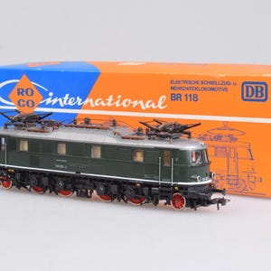 May include: A dark green model electric locomotive with a silver roof and red wheels, positioned in front of its original orange and blue Roco International packaging box labeled with the text Elektrische Schnellzug- u. Mehrzwecklokomotive BR 118.
