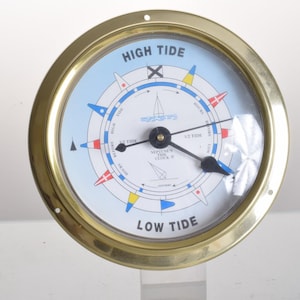 Op de afbeelding: Een ronde getijdenklok met een gepolijste messing rand en een lichtblauwe wijzerplaat. De wijzerplaat bevat de tekst HIGH TIDE en LOW TIDE, samen met nautische vlagmarkeringen, een zeilbootafbeelding en zwarte wijzers voor de getijdenstand.