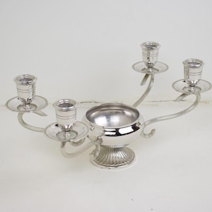 Op de afbeelding: Zilverkleurige kandelaar met vier kaarshouders en een centrale kom. De kaarshouders zijn rond de kom geplaatst op gebogen armen. De kandelaar heeft een decoratieve voet en staat tegen een witte achtergrond.