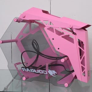 Può includere: Un case per computer rosa con un pannello laterale trasparente. Il case ha un design geometrico con componenti interni visibili e il logo "COUGAR". Il case è realizzato in metallo e plastica.