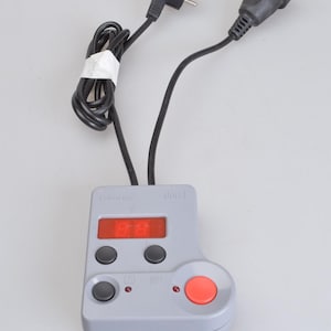 May include: A gray Durst Labotim electronic timer with a digital red display, two black buttons, a smaller black button, and a large red circular button. The device features two black power cords, one with a plug and one with a socket connector.