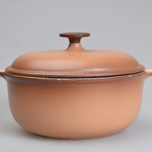 Op de afbeelding: Een ronde, lichtbruine geëmailleerde gietijzeren braadpan met bijpassend deksel. De pan heeft twee zijhandgrepen en een centrale knop op het deksel. De buitenkant heeft een gladde, matte afwerking met zichtbare slijtage langs de bovenrand.
