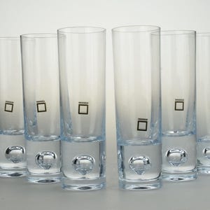 Puede incluir: Un juego de seis vasos de cristal transparente, cada uno con forma cilíndrica y una base gruesa y pesada que contiene una burbuja de aire. Cada vaso está marcado con un pequeño logotipo cuadrado negro en el lateral del cuerpo de cristal.