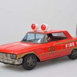 Pode incluir: Um carro de brinquedo de lata vermelho vintage com a inscrição FIRE CHIEF na lateral e no capô. Possui uma antena prateada com ponta rosa, duas luzes vermelhas no teto e um gráfico dourado de machados cruzados nas portas laterais.