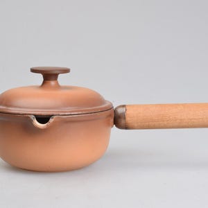 Op de afbeelding: Een matte, terracotta-kleurige keramische steelpan met een bijpassend deksel met een centrale knop. De pan heeft een schenktuit aan de zijkant en een cilindervormig, lichtgekleurd houten handvat bevestigd aan een metalen basis met een oogje.