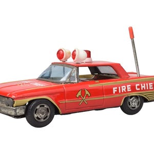 Könnte beinhalten: Ein rotes Spielzeugauto aus Blech mit der Aufschrift FIRE CHIEF an der Seite und auf der Motorhaube. Es verfügt über goldene Verzierungen, zwei rote Dachleuchten, eine silberne Antenne mit roter Spitze und ein gekreuztes Axt-Symbol an der Tür.