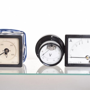 Può includere: Tre strumenti di misura elettrici con cornici nere. A sinistra, un voltmetro quadrato con scala da 0 a 400 V e fili blu. Al centro, un voltmetro rotondo con scala fino a 250 V. A destra, un amperometro quadrato con scala fino a 10 A.