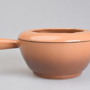 May include: A round ceramic fondue pot with a smooth, glossy finish that transitions from a light peach color at the top rim to a darker, muted brown at the base. It features a single long handle on one side and a small tab handle on the other.
