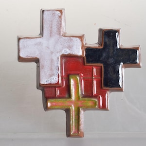 Può includere: Un'opera d'arte da parete in ceramica composta da tre forme a croce sovrapposte. La croce in alto a sinistra è bianca, quella in alto a destra è nera e la croce centrale inferiore è verde lime. Sono sovrapposte a una base rossa a griglia.