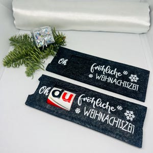 Könnte beinhalten: Zwei dunkelgraue Weihnachtsgeschenkhüllen aus Filz mit weißem Text „Fröhliche Weihnachtszeit“ und „Oh“. Eine Hülle enthält einen Schokoriegel. Eine kleine Geschenkbox und Tannenzweige sind ebenfalls vorhanden.