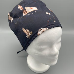 Könnte beinhalten: Eine schwarze OP-Haube mit einem Muster aus Federn, Schriftrollen und Umschlägen. Die Haube wird am Rücken gebunden und auf einem weißen Schaufensterkopf präsentiert. Das Design umfasst braune und weiße Akzente.