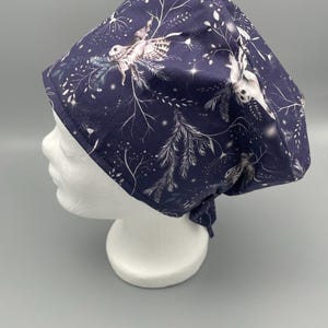 Könnte beinhalten: Eine marineblaue OP-Haube mit einem sich wiederholenden Muster aus weißen Eulen, Zweigen und Sternen. Die Haube ist hinten gebunden und wird auf einem weißen Schaufensterkopf präsentiert. Der Stoff hat eine weiche, matte Oberfläche.