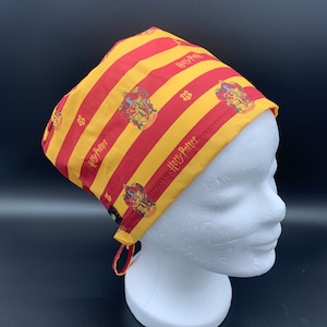 Könnte beinhalten: Eine rot-gelb gestreifte OP-Mütze mit dem Harry-Potter-Logo und -Wappen, das sich über den gesamten Stoff wiederholt. Die Mütze hat einen Bindegurt an der Rückseite.