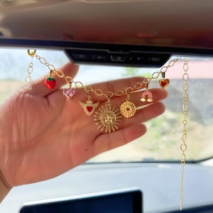 May include: A gold-colored charm chain with various pendants, including a strawberry, hearts, a sun, and a horseshoe. The chain is suspended from a car's rearview mirror. The charms are in red, pink, and gold.