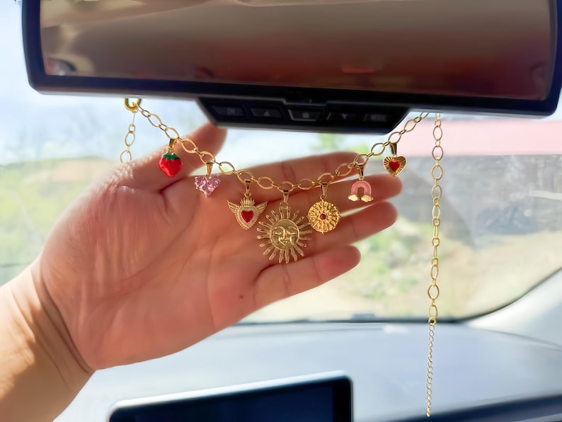 May include: A gold-colored charm chain with various pendants, including a strawberry, hearts, a sun, and a horseshoe. The chain is suspended from a car's rearview mirror. The charms are in red, pink, and gold.