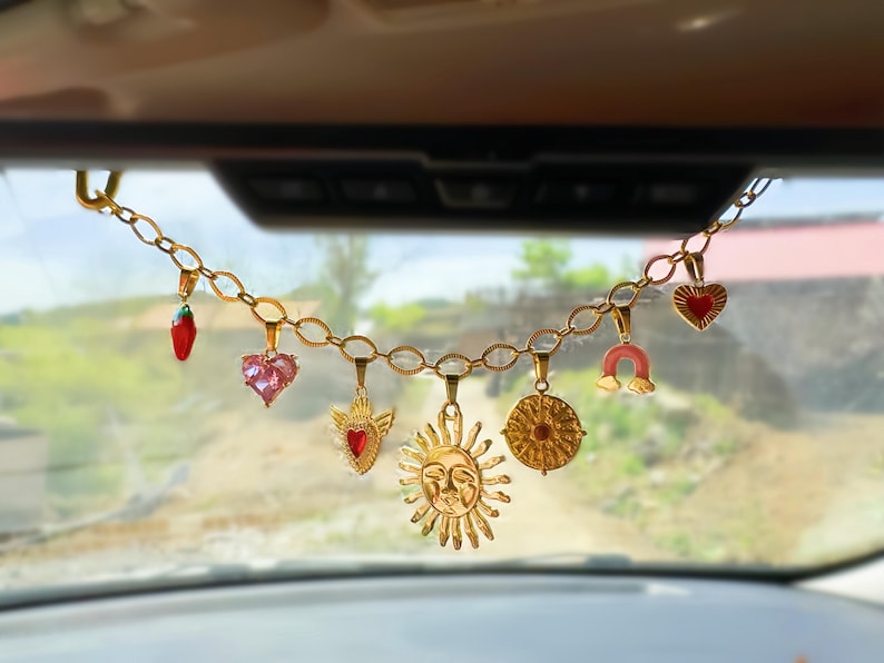 May include: Gold-colored chain with various charms hanging from it. Charms include a red crystal, a pink heart, a sun with a face, a heart with a red center, a horseshoe, and a round charm. The chain hangs in front of a car's windshield.