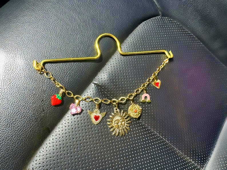 May include: Gold-toned charm bracelet with a variety of pendants, including a strawberry, heart with wings, sun, and horseshoe. The bracelet is attached to a gold-colored safety pin. The charms are in red, pink, and gold.