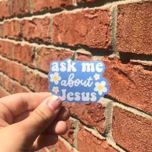 May include: Blue sticker with white daisies and the text "ask me about Jesus" on a brick wall background.