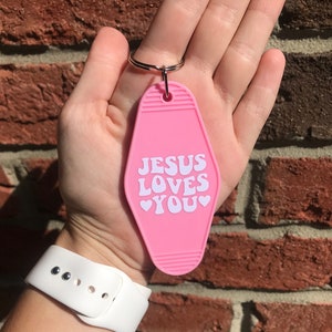 May include: Pink motel key chain with the text "JESUS LOVES YOU" in white lettering with hearts around the word "YOU".
