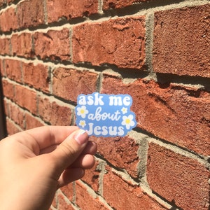 May include: A blue sticker with white flowers and the text "ask me about Jesus" on a brick wall.