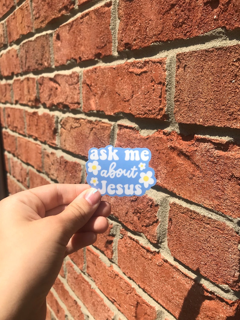 May include: A blue sticker with white flowers and the text "ask me about Jesus" on a brick wall.