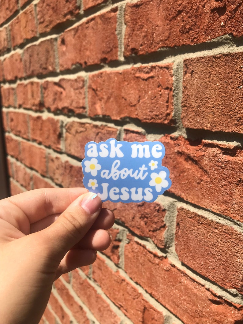 May include: Blue sticker with white daisies and the text "ask me about Jesus" on a brick wall background.