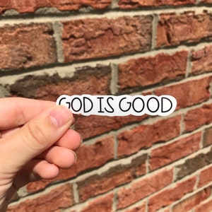 Puede incluir: Una pegatina blanca con texto negro que dice "GOD IS GOOD" sobre un fondo de pared de ladrillo.