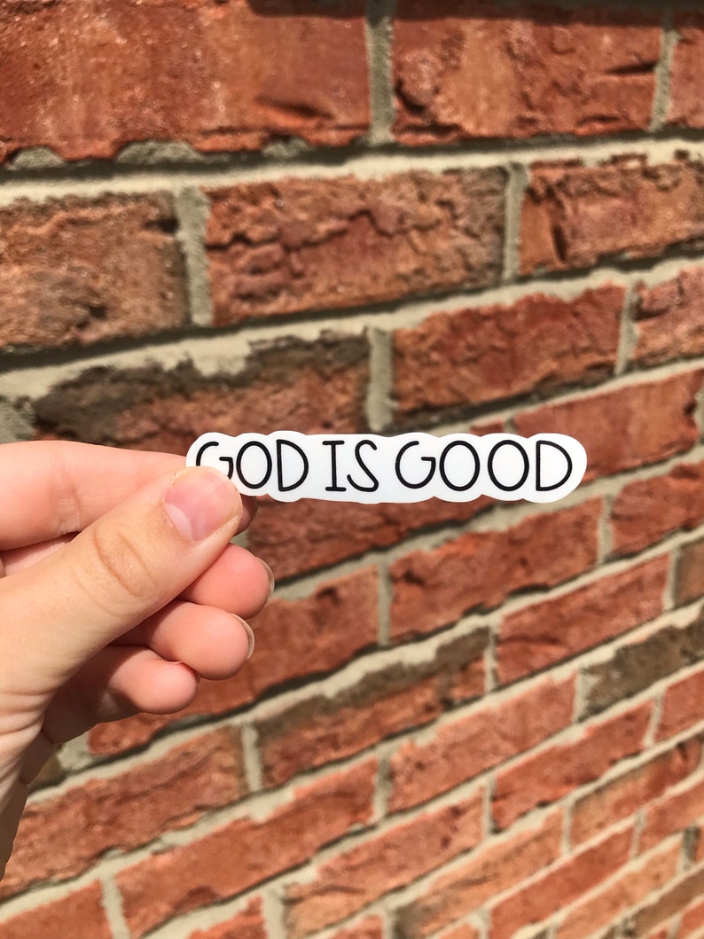 Puede incluir: Una pegatina blanca con texto negro que dice "GOD IS GOOD" sobre un fondo de pared de ladrillo.