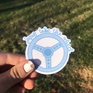 May include: A light blue sticker featuring a steering wheel design with the words "Jesus take the wheel" in a cursive font. The sticker has a white background and a light blue border.
