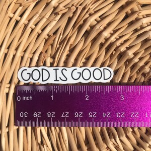 Puede incluir: Pegatina blanca con contorno negro y el texto "GOD IS GOOD" sobre un fondo de mimbre tejido. Una regla morada con marcas en pulgadas est&aacute; debajo de la pegatina.